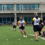 Pittsburgh Steelers quarterback Will Howard (18) runs with the football during an OTA drill at the UPMC Rooney Sports Complex, with a teammate observing and the team facility visible in the background.
