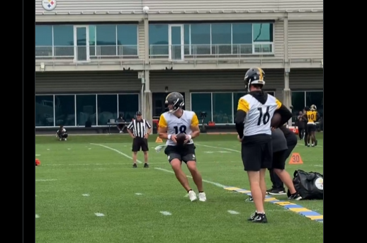 Pittsburgh Steelers quarterback Will Howard (18) runs with the football during an OTA drill at the UPMC Rooney Sports Complex, with a teammate observing and the team facility visible in the background.